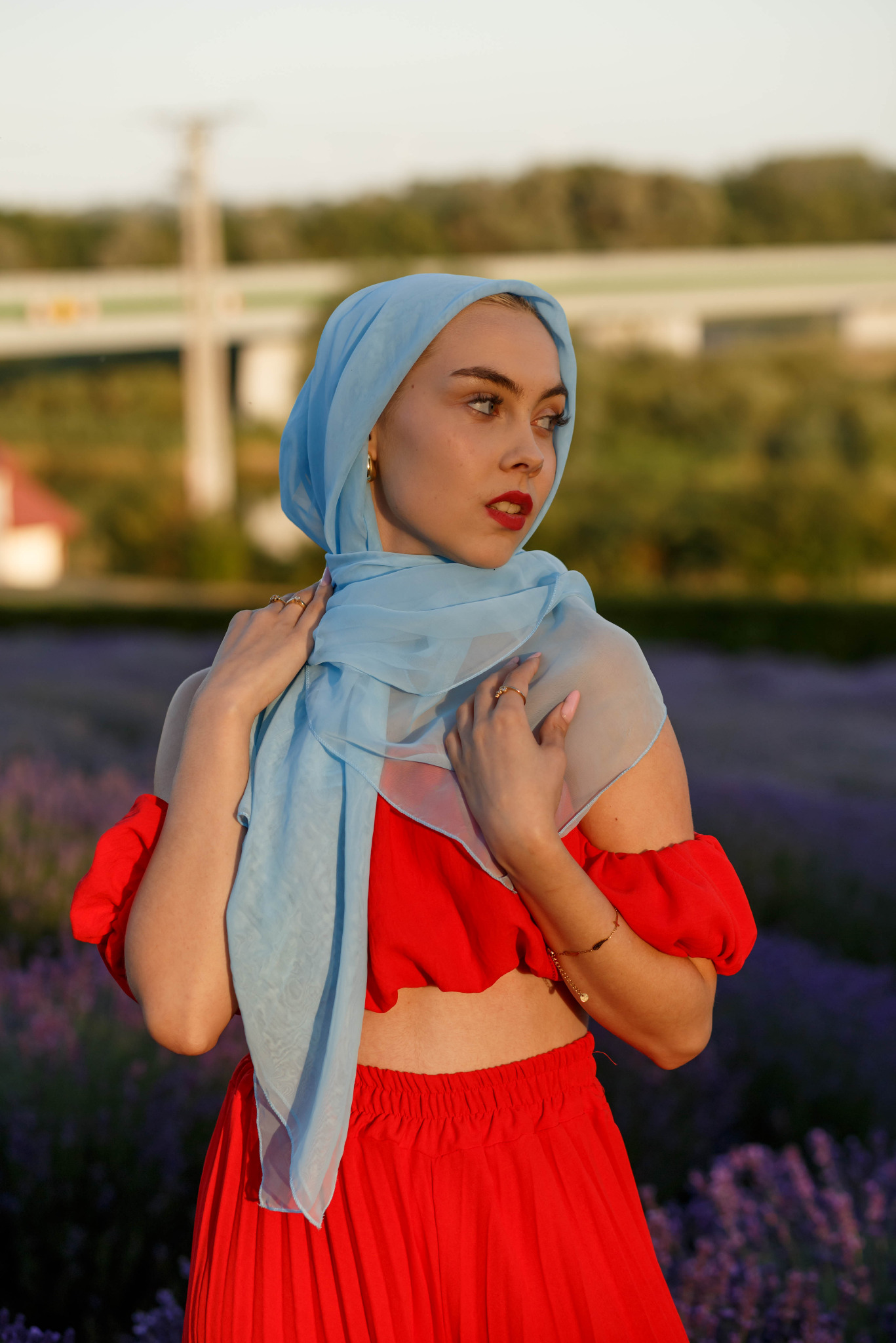 Portret kobiety w czerwonym stroju i błękitnym hidżabie na tle pola lawendy. Ujęcie z dołu, delikatne światło podkreśla urodę modelki. W tle widoczna zieleń i fragment zabudowań.