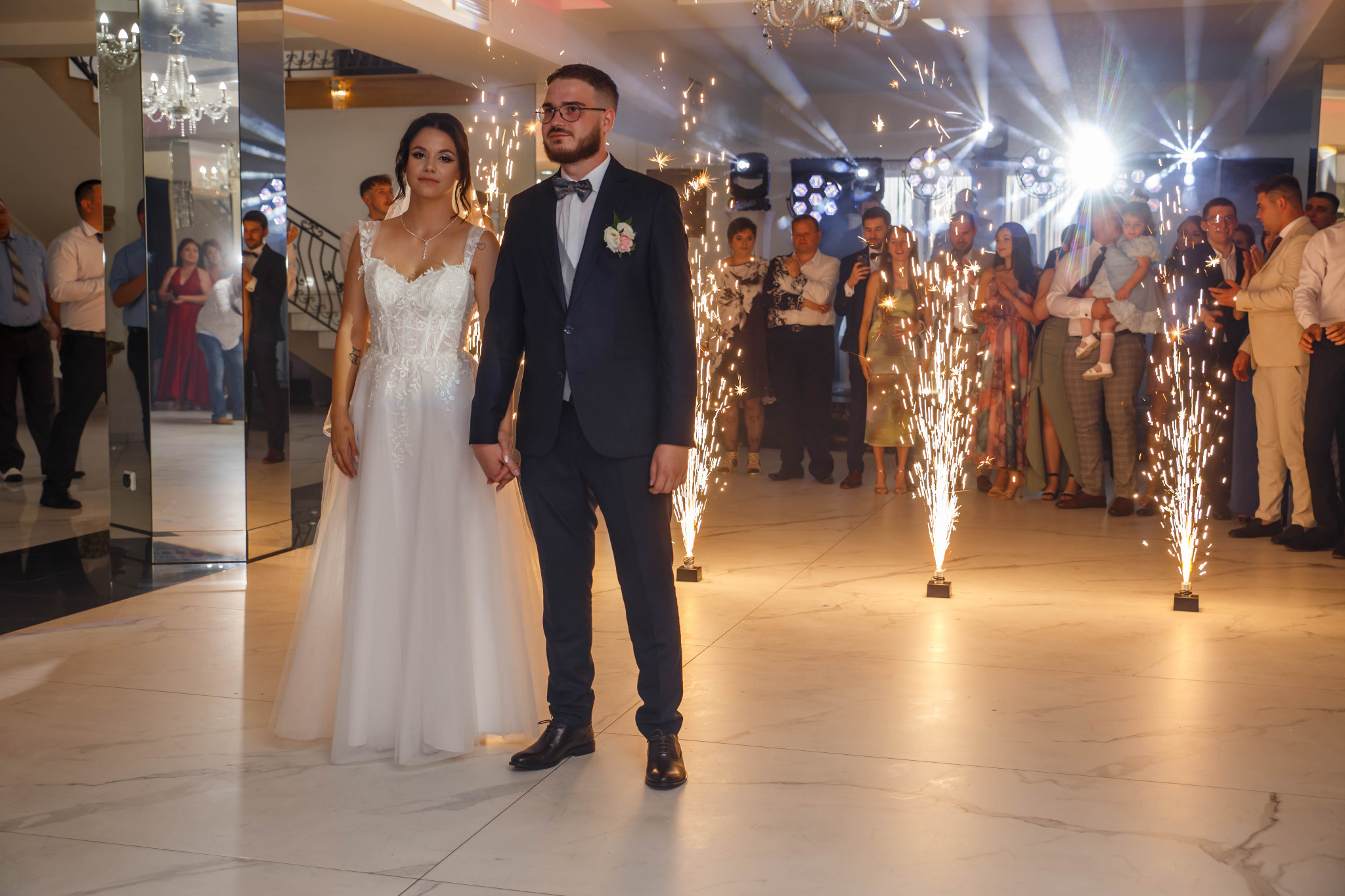 Para młoda trzymająca się za ręce na tle gości weselnych i fontanny iskier. Panna młoda w białej sukni, pan młody w garniturze. Ujęcie z uroczystości weselnej.