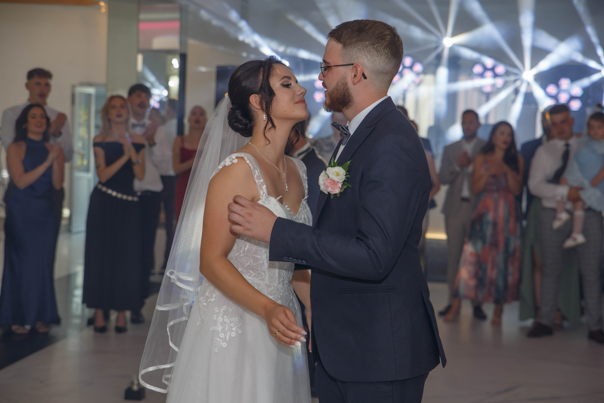 Ujęcie pary młodej podczas pierwszego tańca, w tle goście weselni i dekoracje świetlne. Pan młody w ciemnym garniturze, panna młoda w białej sukni z welonem.