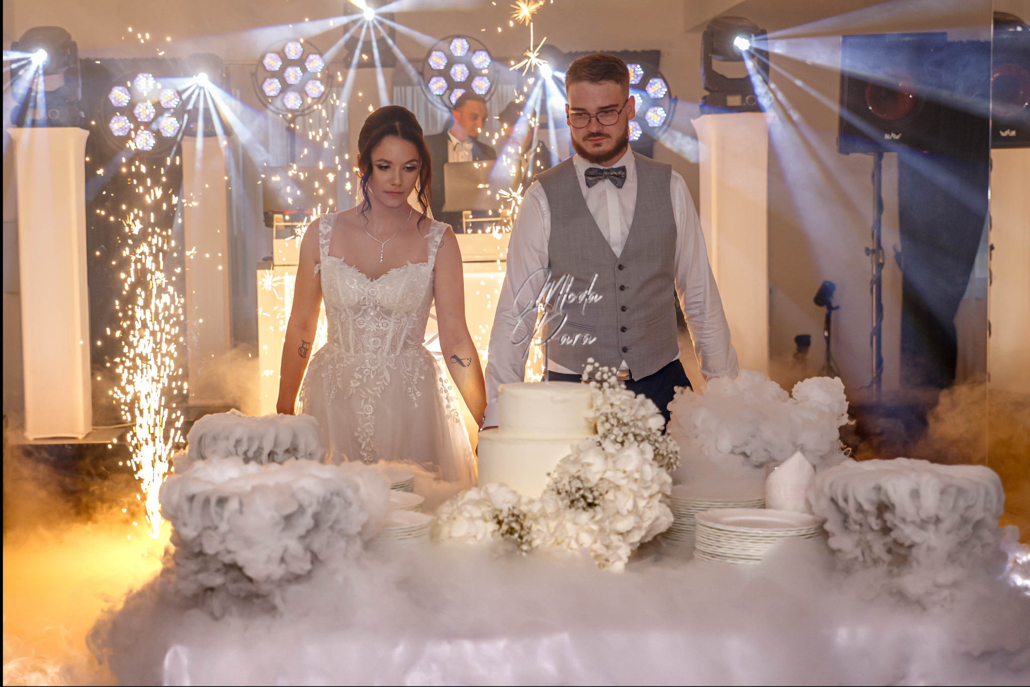 Uroczyste krojenie tortu weselnego przez parę młodą, w otoczeniu sztucznych ogni i dekoracji z dymu. W tle DJ za konsoletą i oświetlenie sceniczne.