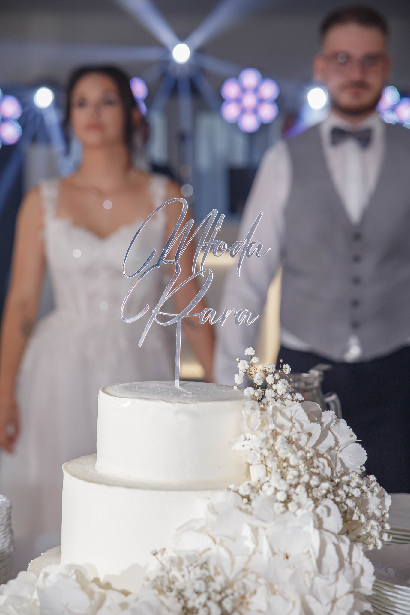 Dwupoziomowy tort weselny z kwiatami i srebrnym napisem 'Młoda Para', na rozmytym tle para młoda i dekoracje świetlne.