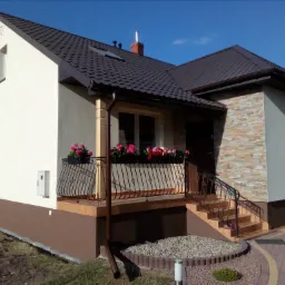 Dwukondygnacyjny dom z ciemnobrązowym dachem, jasną elewacją i kamienną okładziną fragmentu ściany, ozdobiony kwiatami w skrzynkach balkonowych i wykończony brązowym cokołem.