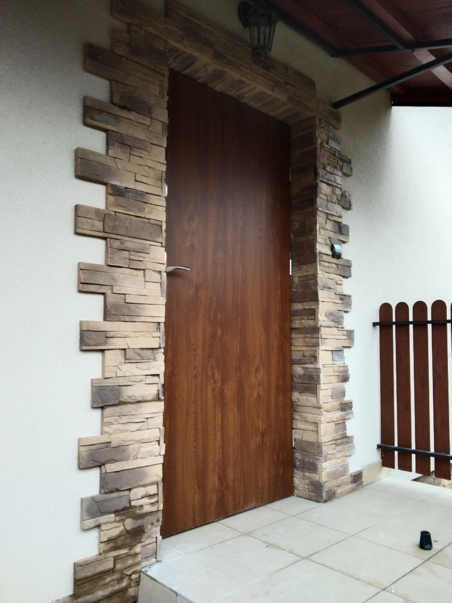 Wejście do domu z drewnianymi drzwiami obramowane dekoracyjną elewacją z kamienia elewacyjnego w odcieniach beżu i brązu, z widoczną lampą zewnętrzną nad wejściem i fragmentem drewnianego płotu.