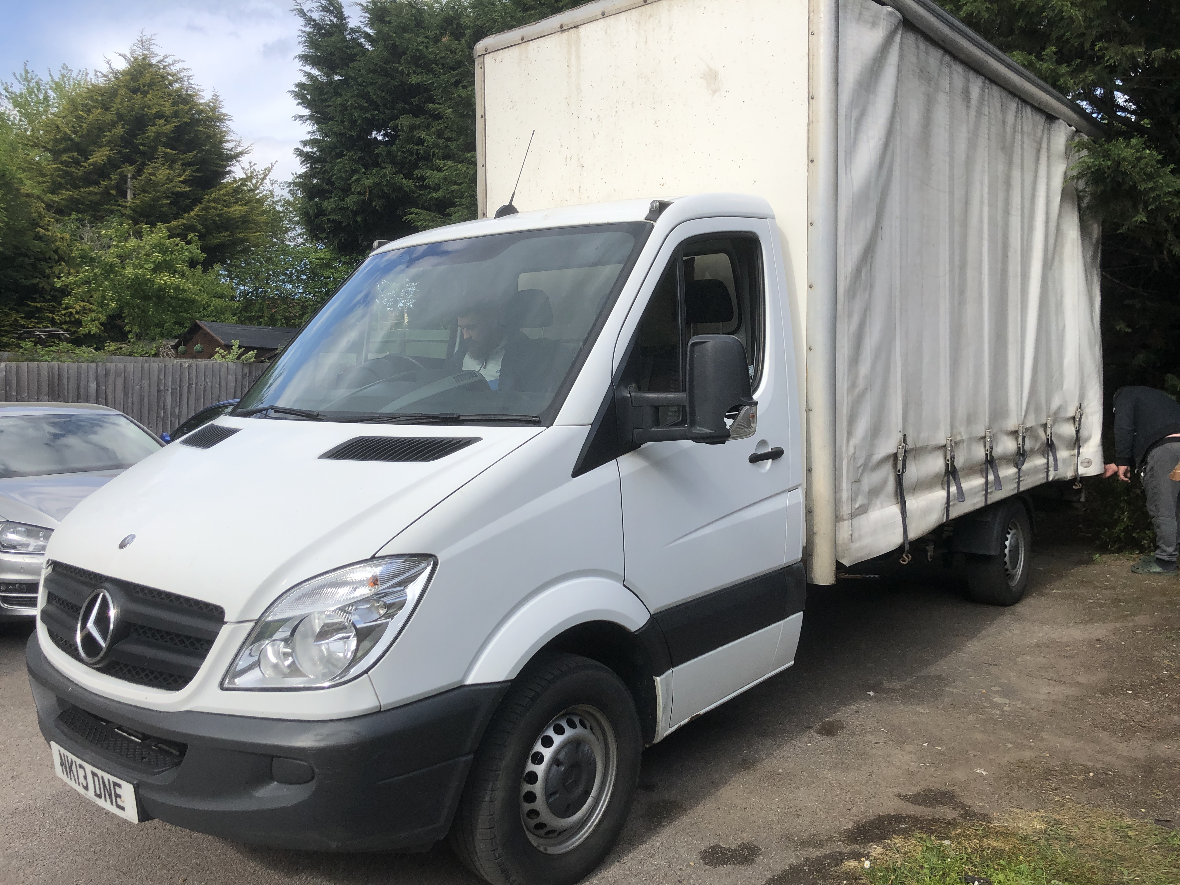 Biały samochód dostawczy Mercedes-Benz Sprinter z plandeką, zaparkowany na podwórku, z widocznym kierowcą i osobą przy plandece z tyłu, tablica rejestracyjna 'NK13 ONE'.