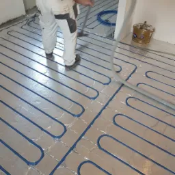 Instalacja ogrzewania podłogowego: niebieskie rurki ułożone na srebrnej folii izolacyjnej, pracownik z odkurzaczem przemysłowym usuwa zanieczyszczenia przed wylaniem wylewki.