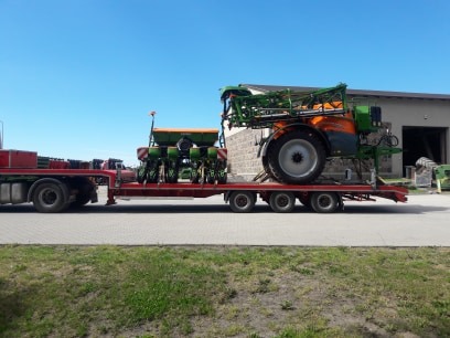Niskopodwoziowa platforma transportowa przewożąca siewnik punktowy i opryskiwacz polowy marki Amazone na tle budynku gospodarczego w słoneczny dzień.