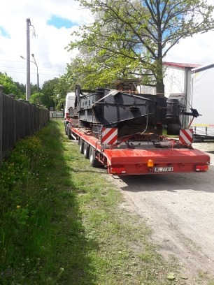 Czerwona naczepa niskopodwoziowa z czarną konstrukcją stalową, zabezpieczona pasami, widoczna z tyłu, zaparkowana na poboczu z zieloną trawą i drewnianym płotem w tle.