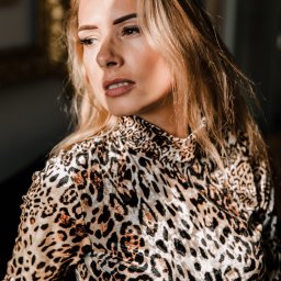 Marika Małek - Portret kobiety w bluzce w panterkę, z delikatnym makijażem, spoglądającej w bok, oświetlonej częściowo promieniami słońca.
