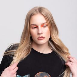 Marika Małek - Portret modelki w czarnym T-shircie z nadrukiem motyla i planety, z pomarańczowym makijażem oczu, na jasnym tle. Dłonie w jasnych włosach.