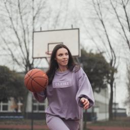 Marika Małek - Kobieta w liliowym dresie z logo 'Claire' na bluzie, trzyma piłkę do koszykówki na tle boiska.