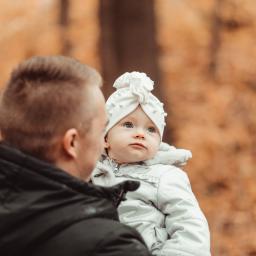 Marika Małek - Ojciec trzyma na rękach niemowlę w białej kurtce i czapce z ozdobami, jesienne tło w Toruniu.
