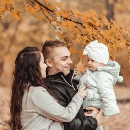 Marika Małek - Jesienna sesja rodzinna: Uśmiechnięta para trzyma na rękach niemowlę w białym kombinezonie i czapce, z opadającymi żółtymi liśćmi w tle.