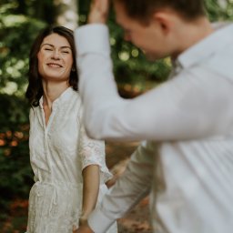 Marika Małek - Uśmiechnięta kobieta w koronkowej sukience spogląda w lewo, trzymając za rękę lekko rozmazanego mężczyznę w białej koszuli na tle zieleni drzew.