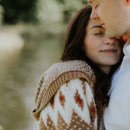Marika Małek - Zbliżenie na parę zakochanych: brunetka w swetrze z etnicznym wzorem i mężczyzna w białej koszuli, dotykający się czołami na tle rozmytego jeziora i zieleni.