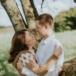 Marika Małek - Para narzeczonych przytulona do drzewa, kobieta w swetrze z wzorem, mężczyzna w białej koszuli, oboje z zamkniętymi oczami i uśmiechem.