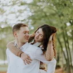 Marika Małek - Młoda para: mężczyzna trzyma kobietę na barana w parku, ona opiera głowę na jego ramieniu, uśmiechając się z zamkniętymi oczami, w tle rozmyte drzewa.