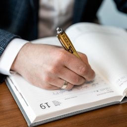 Marika Małek - Osoba w marynarce wpisuje coś do otwartego kalendarza długopisem z logo firmy Walaszko BHP, widoczny dzień tygodnia i numer 19.