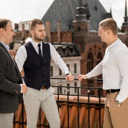 Marika Małek - Trzech mężczyzn w eleganckich strojach rozmawia na tle zabytkowej architektury z czerwonej cegły, widocznej z balkonu z pomarańczową balustradą.