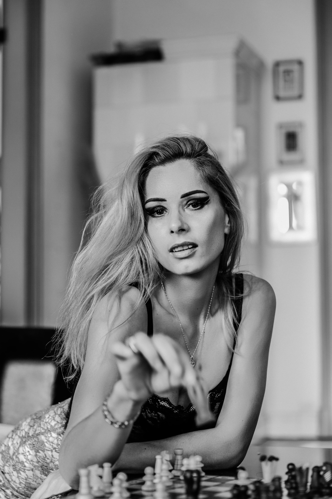 Czarno-białe ujęcie kobiety z blond włosami, grającej w szachy, z wyrazistym makijażem oczu i delikatnym naszyjnikiem, siedzącej przy szachownicy w jasnym pomieszczeniu.