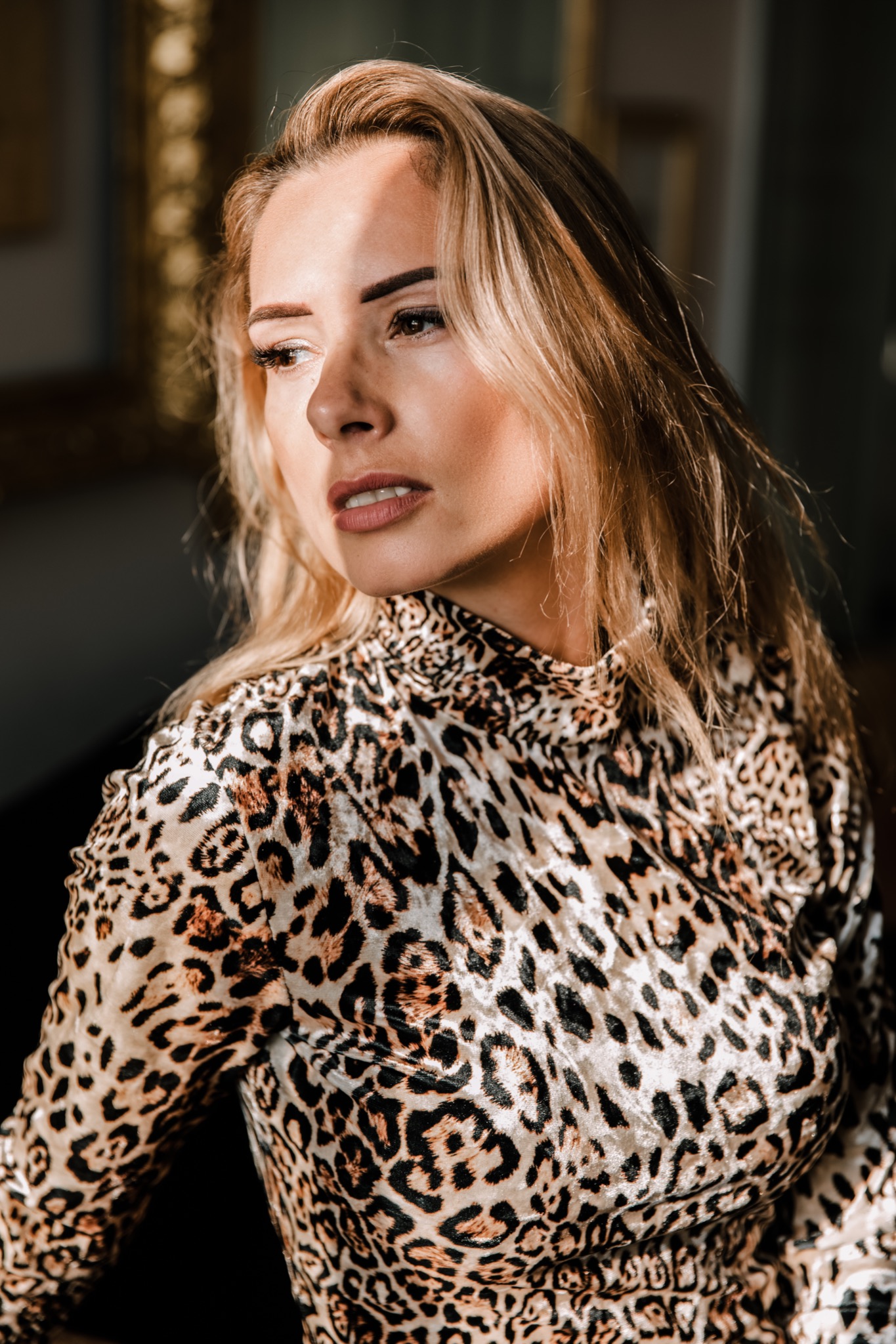 Portret kobiety w bluzce w panterkę, z delikatnym makijażem, spoglądającej w bok, oświetlonej częściowo promieniami słońca.