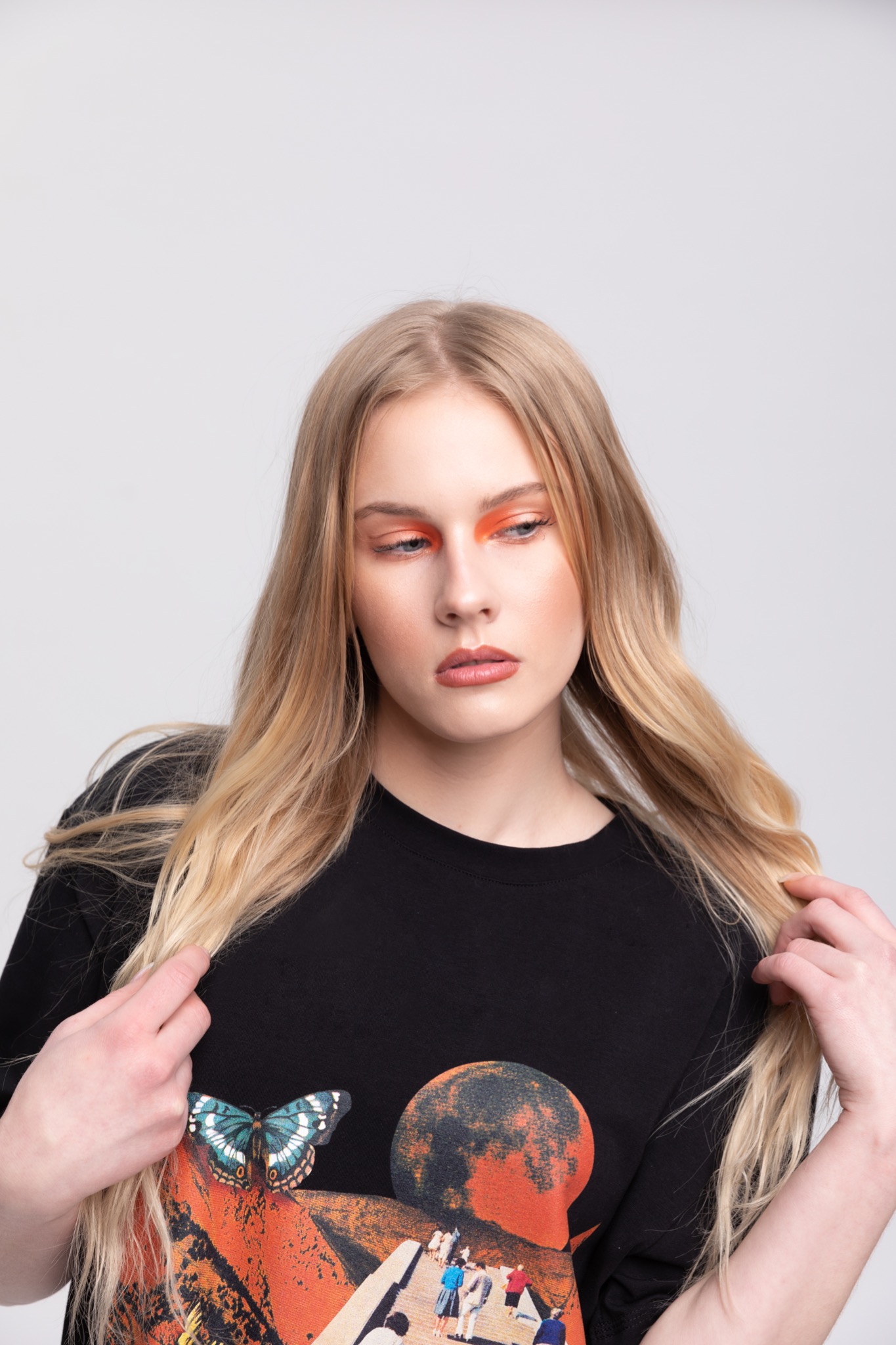 Portret modelki w czarnym T-shircie z nadrukiem motyla i planety, z pomarańczowym makijażem oczu, na jasnym tle. Dłonie w jasnych włosach.