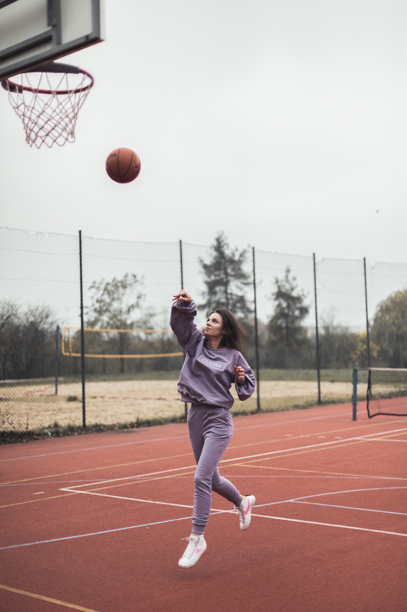 Kobieta w liliowym dresie marki Claire rzuca piłkę do kosza na czerwonym boisku sportowym.