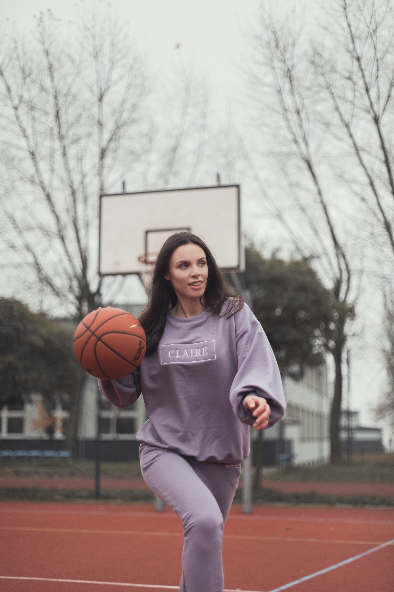 Kobieta w liliowym dresie z logo 'Claire' na bluzie, trzyma piłkę do koszykówki na tle boiska.