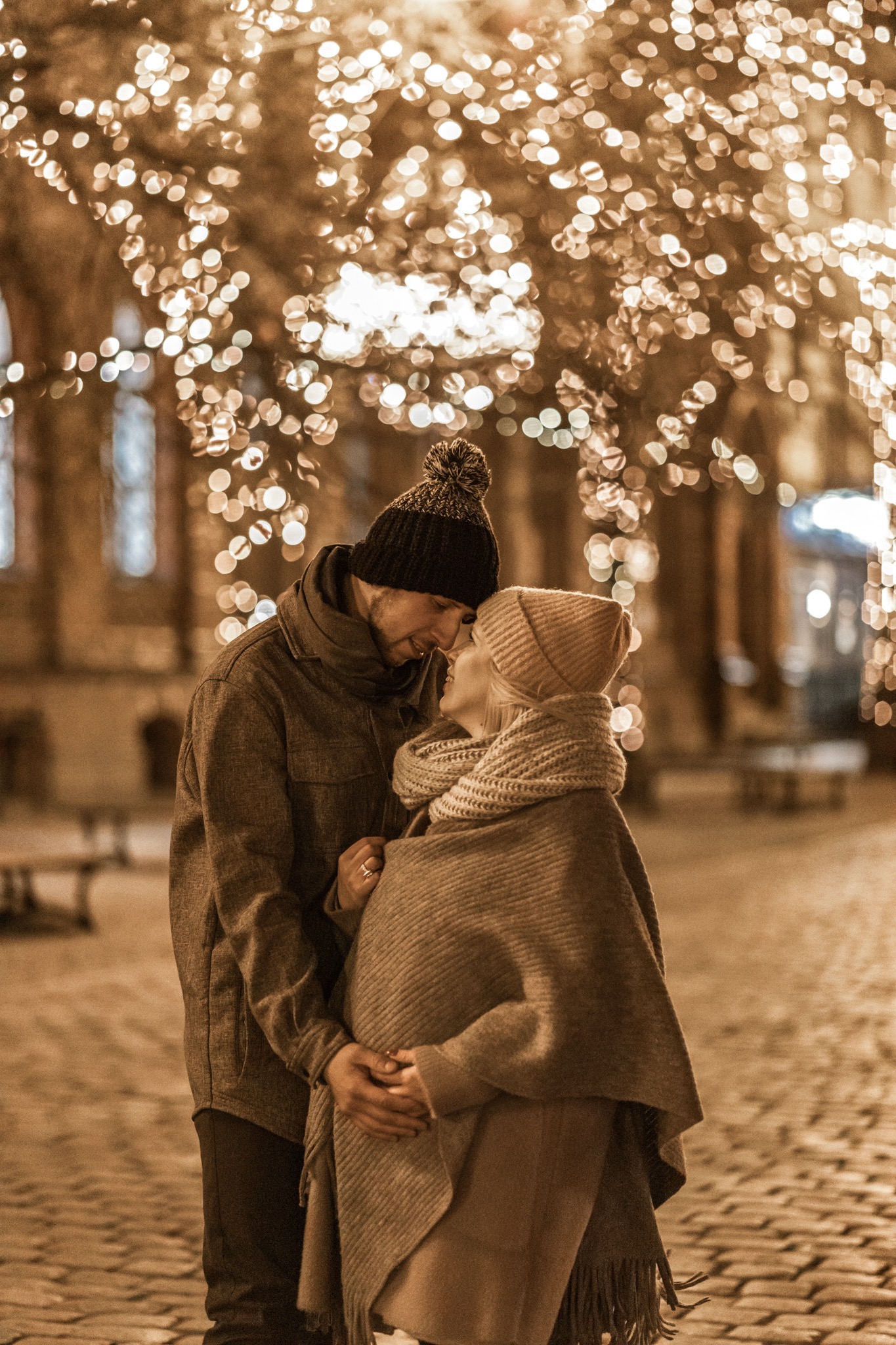 Przyszła mama w beżowej czapce i ponczo, wtulona w partnera pod rozświetlonymi drzewami, delikatne ujęcie z bliska.