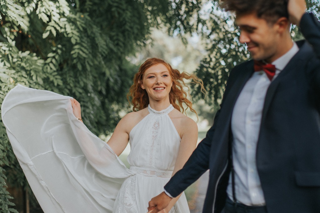Uśmiechnięta, ruda kobieta w białej sukni z koronkowymi detalami i zwiewnym szalem, trzymająca za rękę mężczyznę w granatowym garniturze i bordowej muszce, na tle zielonych drzew.