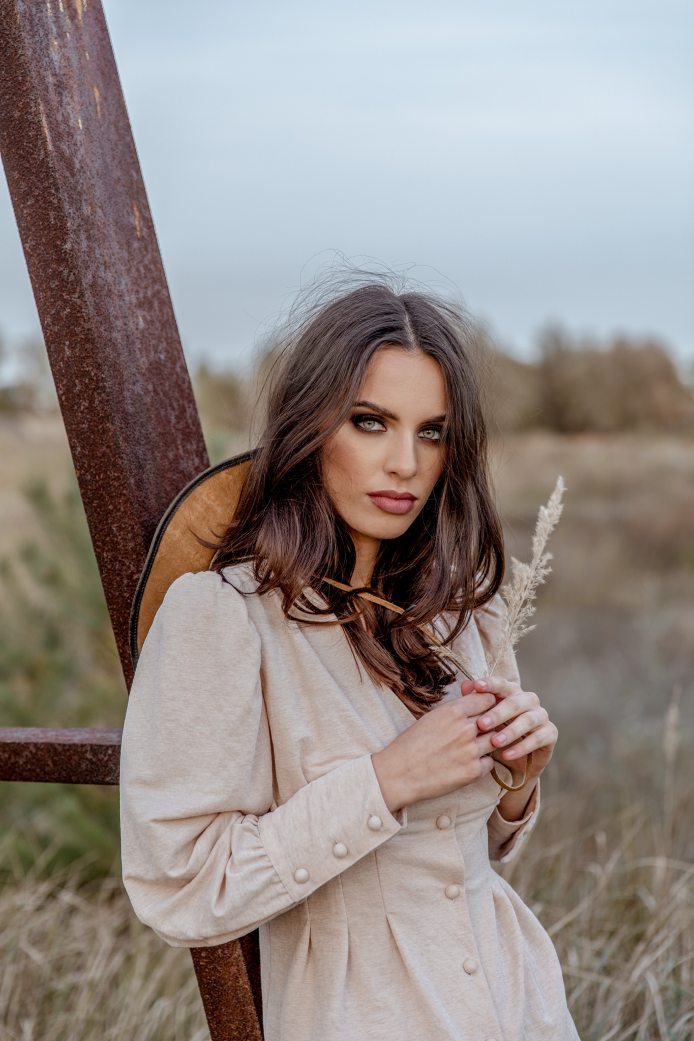 Portret kobiety o przenikliwym spojrzeniu, w beżowej sukience z bufiastymi rękawami, trzymającej źdźbło trawy na tle zardzewiałej konstrukcji i jesiennego krajobrazu.