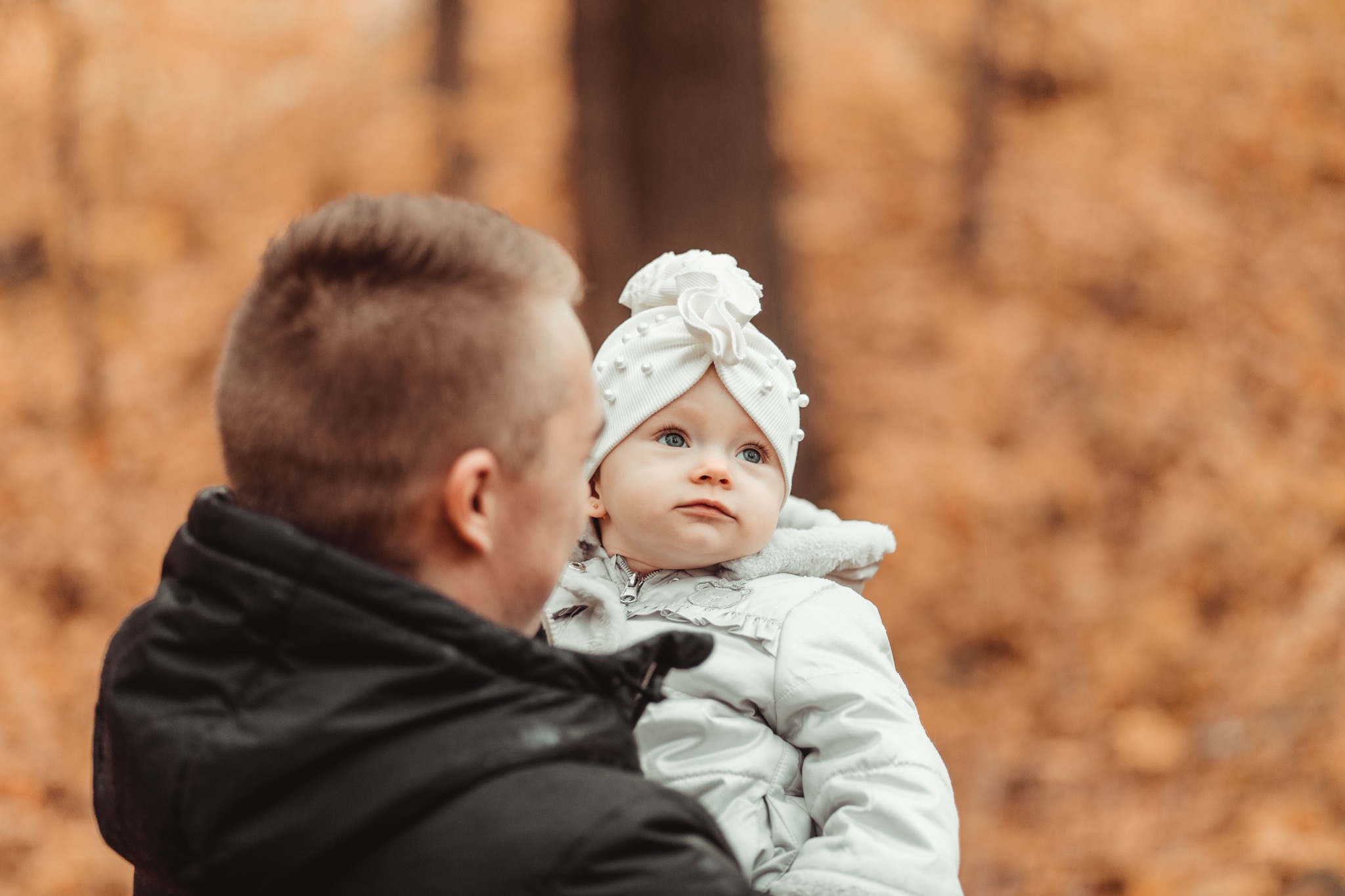 Ojciec trzyma na rękach niemowlę w białej kurtce i czapce z ozdobami, jesienne tło w Toruniu.