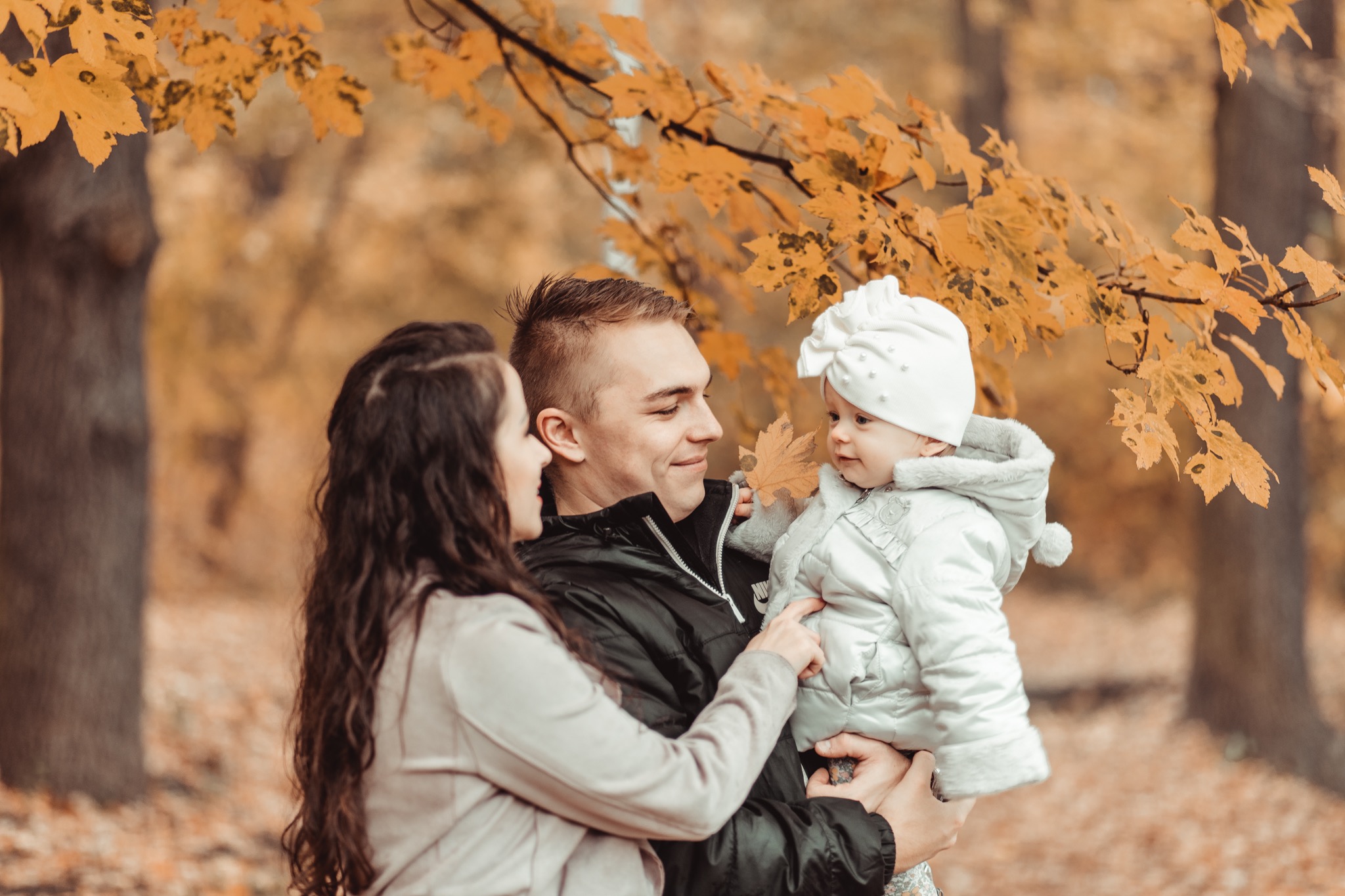 Jesienna sesja rodzinna: Uśmiechnięta para trzyma na rękach niemowlę w białym kombinezonie i czapce, z opadającymi żółtymi liśćmi w tle.