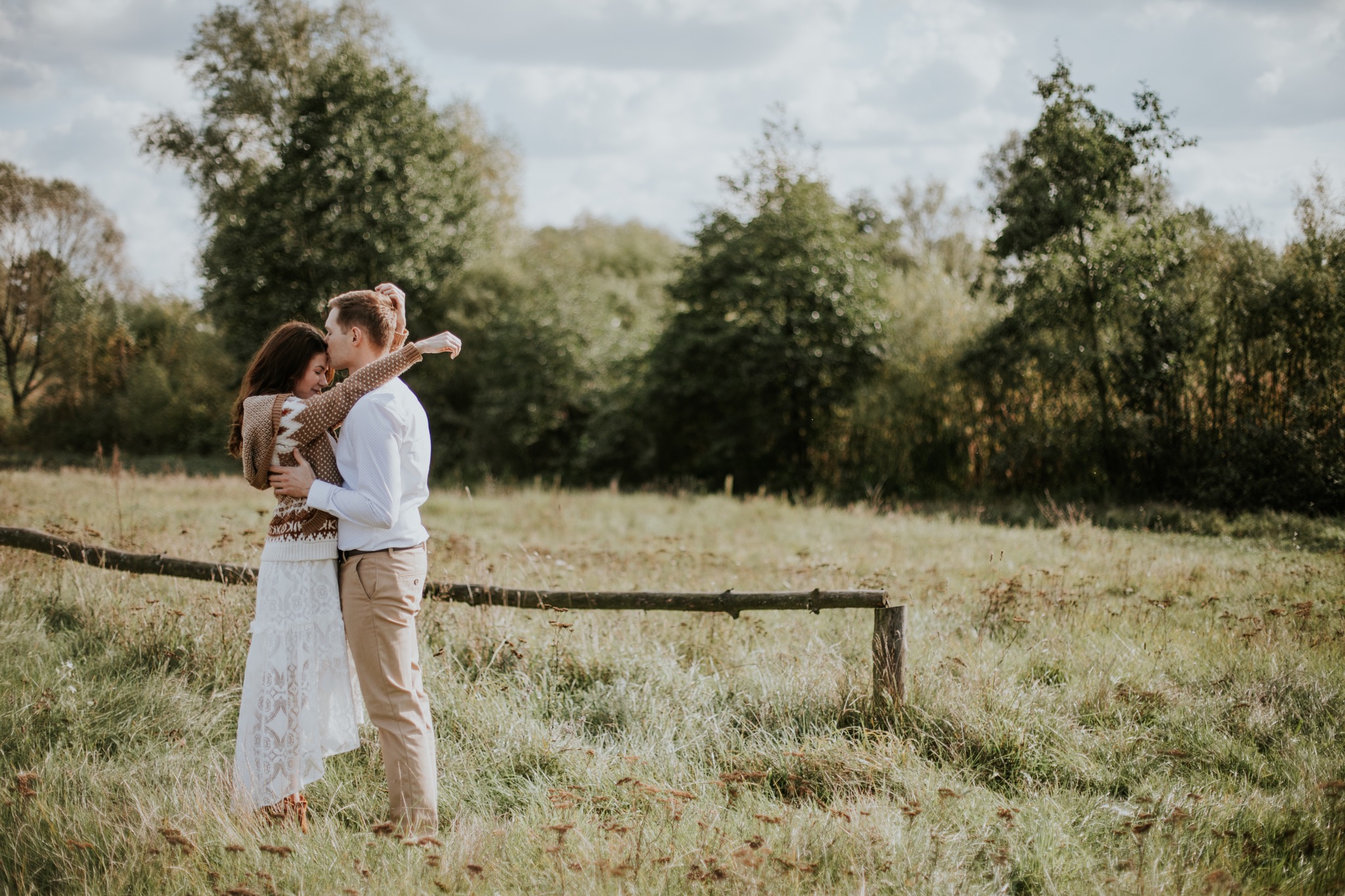 Para obejmująca się na łące w pobliżu drewnianego płotku, kobieta w koronkowej sukience i swetrze w etniczne wzory, mężczyzna w białej koszuli i beżowych spodniach, w tle drzewa i pochmurne niebo.