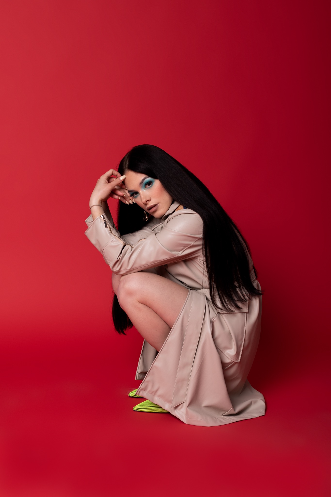 Kobieta w beżowym płaszczu i neonowych butach pozuje na czerwonym tle, z mocnym makijażem oczu w odcieniach błękitu i brokatu, prezentując kolekcję SS21.