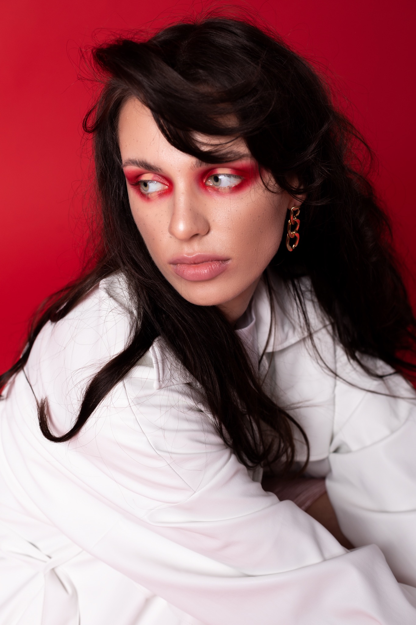 Portret kobiety z ciemnymi włosami i wyrazistym, czerwonym makijażem oczu, pozującej na tle czerwonego tła w białej, oversizowej koszuli i złotych kolczykach.