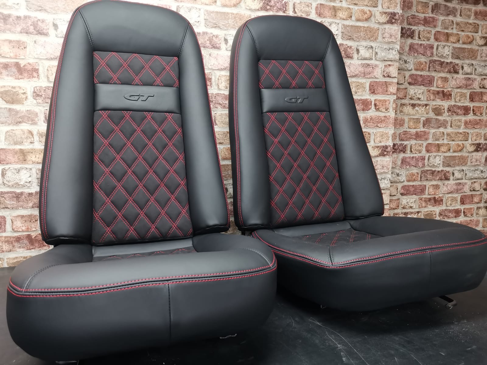 Dwa odnowione, czarne fotele samochodowe z czerwonym przeszyciem w romby, z logo GT, na tle ceglanej ściany.