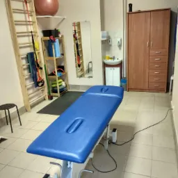 Gabinet fizjoterapii z niebieskim stołem do masażu, drabinką gimnastyczną, piłką rehabilitacyjną i szafką na tle beżowych ścian i kafelkowej podłogi.