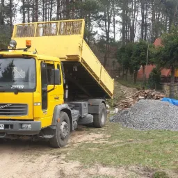 Żółta ciężarówka wywrotka Volvo z podniesioną skrzynią ładunkową stoi na placu budowy obok dwóch pryzm szarego kruszywa, w tle widoczny wykop, stosy drewna i las