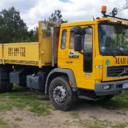 Żółta ciężarówka Volvo FL10 z numerem telefonu na skrzyni ładunkowej firmy MAR-KAR, zaparkowana na trawiastym terenie z widocznym stosem gruzu i betonowym ogrodzeniem w tle.