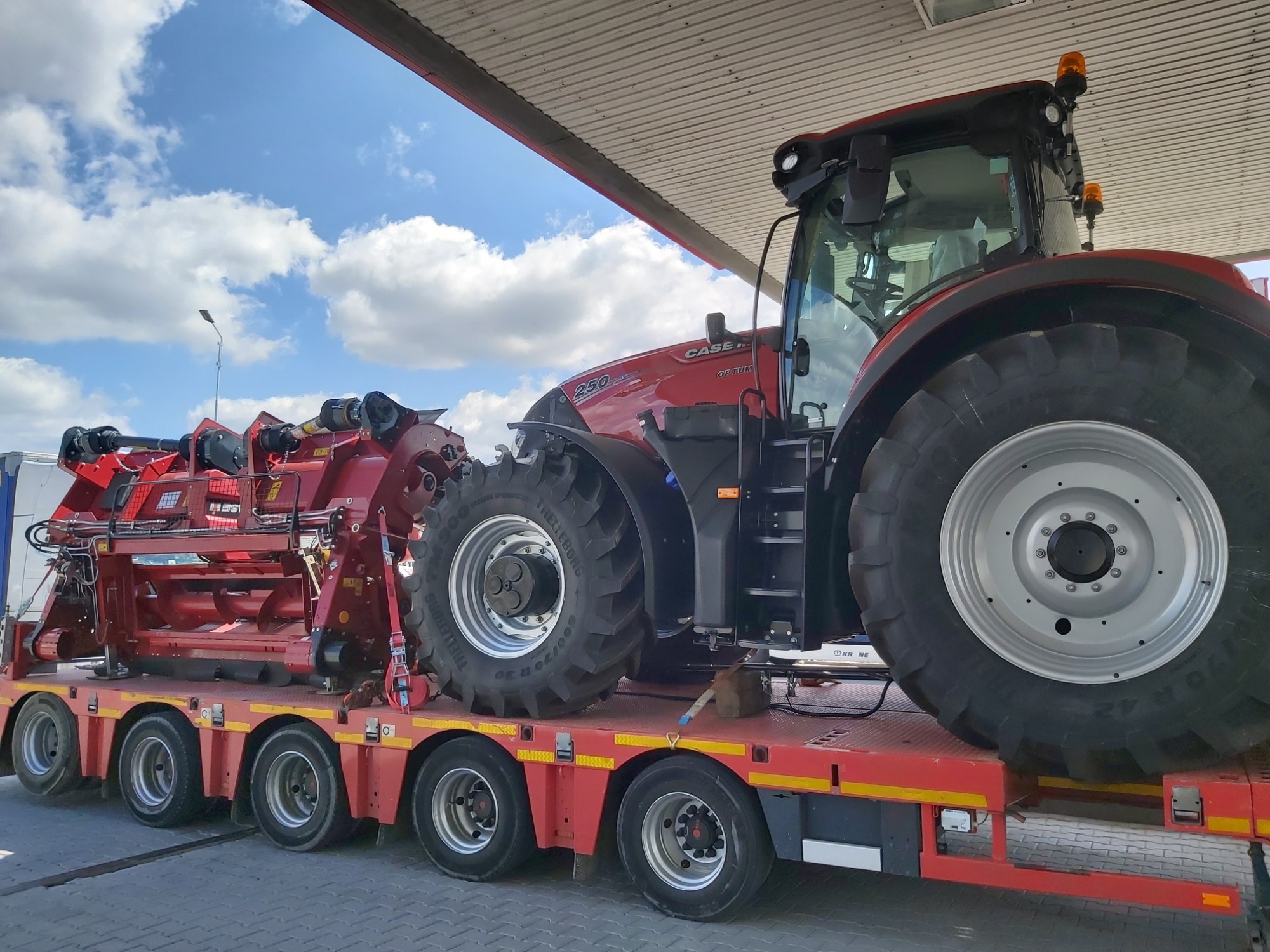 Czerwony ciągnik Case IH Optum 250 na niskopodwoziowej platformie transportowej z zamontowanym czerwonym agregatem uprawowym, widoczne duże opony i błękitne niebo z chmurami.