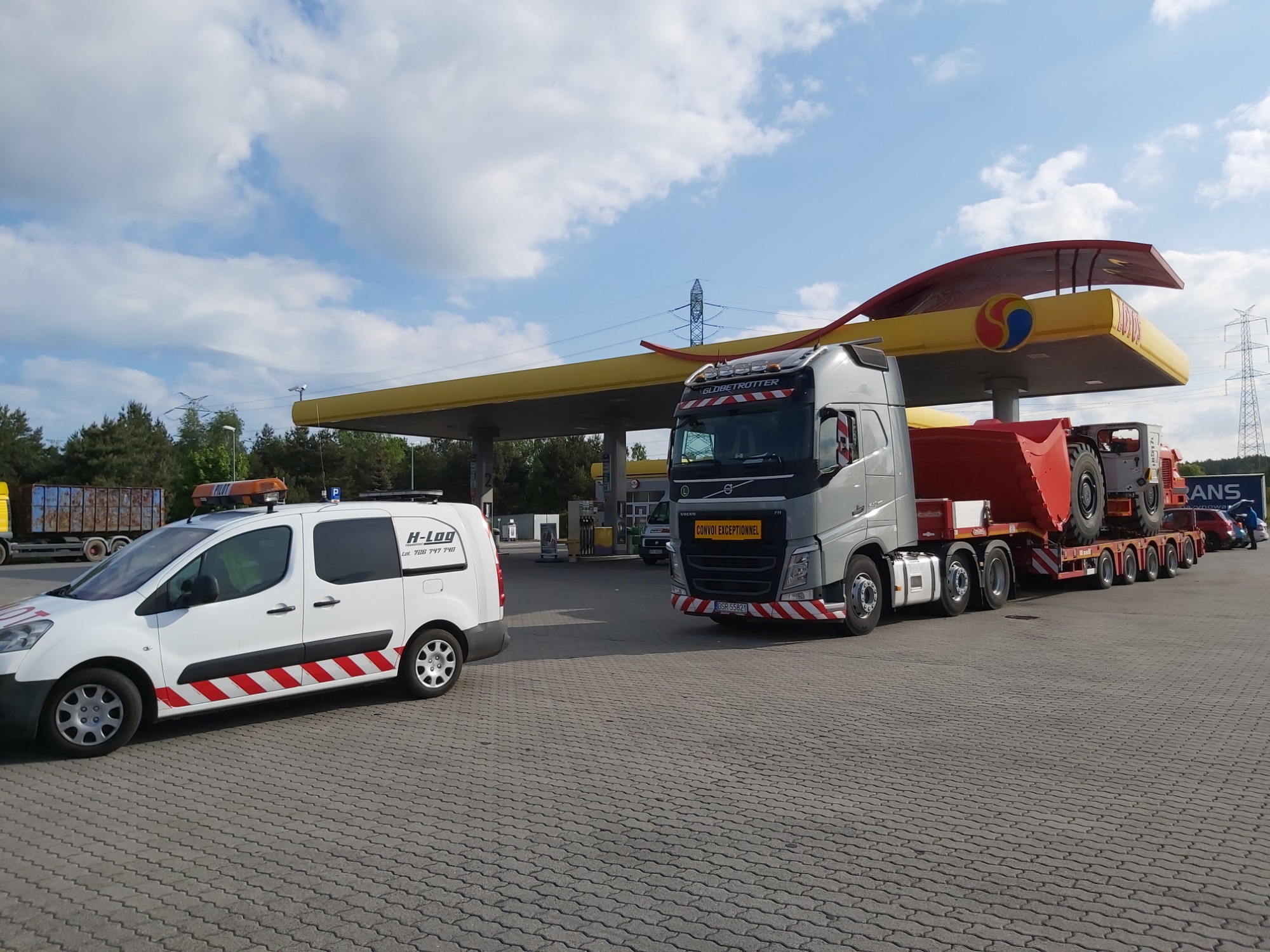 Ciężarówka z naczepą przewożąca ponadgabarytowy ładunek (maszyna budowlana) na stacji benzynowej, obok zaparkowany pojazd pilotujący z oznaczeniami ostrzegawczymi.