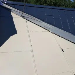 Fragment dachu z blachy trapezowej w kolorze grafitowym oraz fragment ocieplonego i pomalowanego poddasza w jasnym odcieniu beżu, widoczne łączenia płyt i przewód wystający z dachu.