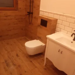 Nowoczesna łazienka z drewnopodobnymi płytkami na ścianach i podłodze, biała umywalka z szafką w stylu retro oraz podwieszana toaleta.