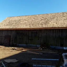 Zrekonstruowana zabytkowa stodoła z nowym dachem pokrytym gontem kaszubskim, widok na drewnianą konstrukcję i jasne drewniane gonty na tle błękitnego nieba.