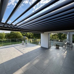 Elkano - Nowoczesny taras z szarych płytek, zadaszony metalową pergolą z regulowanymi lamelami. Szklane balustrady, rośliny w donicach i meble ogrodowe dopełniają aranżację.