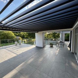 Elkano - Nowoczesny taras z szarą, dużą płytką, zadaszony metalową pergolą z widokiem na zieleń. Szklane balustrady, minimalistyczne donice.