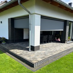 Elkano - Nowoczesny taras z szarej kostki brukowej, obrzeża z ciemnych kostek, częściowo zasłonięty roletą zewnętrzną. Widok na meble ogrodowe i fragment trawnika.