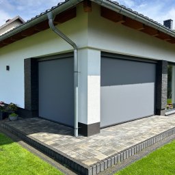 Elkano - Szare rolety zewnętrzne na elewacji budynku z kostką brukową i zielonym trawnikiem w słoneczny dzień. Widoczny fragment dachu z rynną.