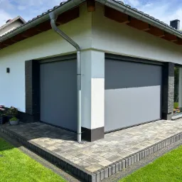 Szare rolety zewnętrzne na elewacji budynku z kostką brukową i zielonym trawnikiem w słoneczny dzień. Widoczny fragment dachu z rynną.