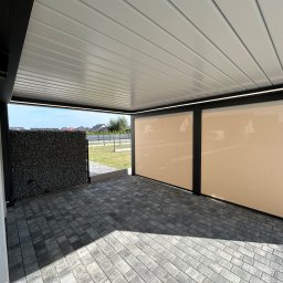 Elkano - Zadaszony taras z szarą kostką brukową i beżowymi roletami bocznymi. Widoczny fragment gabionu z kamienia. Oświetlenie LED pod dachem.